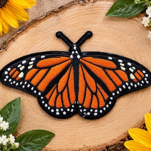 May include: An embroidered butterfly patch featuring vibrant orange wings with black veins and borders, accented by small white dots along the edges. The design includes two black antennae extending from the top center of the butterfly body.