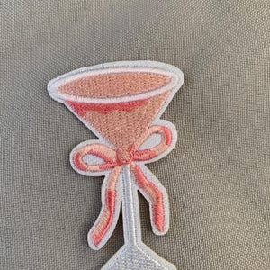 May include: A pink and white embroidered patch of a martini glass with a pink bow.