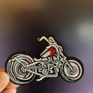 May include: White embroidered patch of a skeleton riding a motorcycle. The motorcycle is black with red accents. The patch is on a black background.