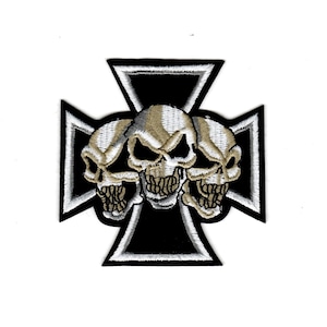 May include: Embroidered patch featuring three skulls in shades of tan and gray, centered on a black and white cross design. The patch has a detailed, textured appearance, suitable for jackets or bags.