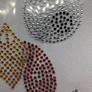 May include: Close-up of a design featuring sparkling rhinestones in various shapes and colors. A large circle is made of silver rhinestones with a black center. Other shapes include a gold crown and a red teardrop.
