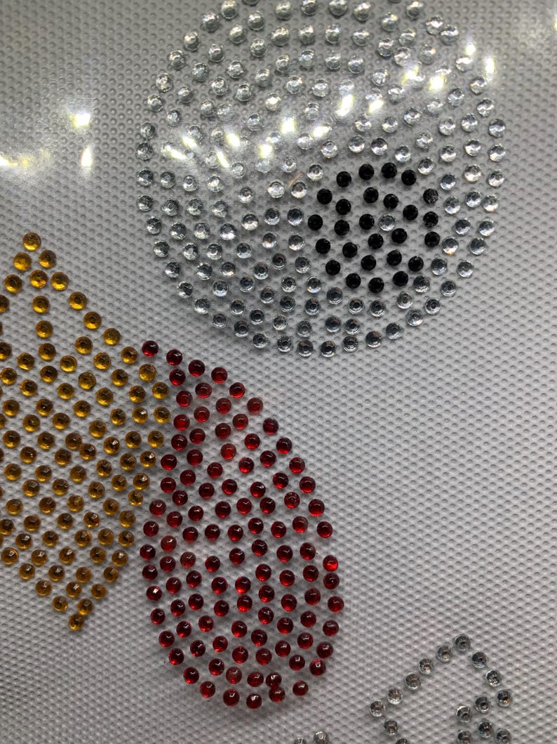 May include: Close-up of a design featuring sparkling rhinestones in various shapes and colors. A large circle is made of silver rhinestones with a black center. Other shapes include a gold crown and a red teardrop.