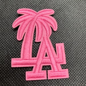 May include: A pink embroidered patch featuring the letters "LA" with a palm tree design above. The patch is a vibrant shade of pink and is set against a textured black background.