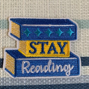 May include: Embroidered patch featuring two stacked books in blue and yellow. The top book has teal star accents. The patch reads "STAY Reading" in white and yellow lettering, promoting literacy and reading.