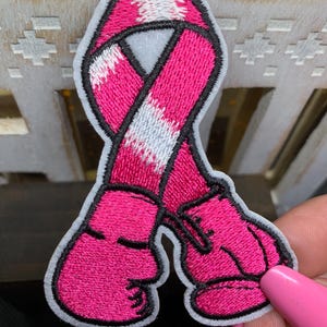 May include: An embroidered patch featuring a pink awareness ribbon intertwined with two bright pink boxing gloves. The design is outlined in black thread with a white border, showcasing detailed stitching throughout the fabric surface.