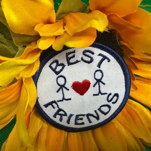 May include: A close-up of a bright yellow sunflower with a circular patch at its center. The patch has the words "BEST FRIENDS" in black lettering around the edge. Two stick figures are on either side of a red heart.