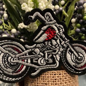 Puede incluir: Parche bordado con un dise&ntilde;o de motocicleta esquel&eacute;tica. El parche tiene un borde negro con detalles blancos y rojos, que muestran la estructura esquel&eacute;tica de la motocicleta. El dise&ntilde;o est&aacute; sobre un fondo de flores artificiales.