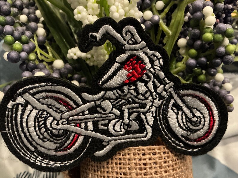 Puede incluir: Parche bordado con un dise&ntilde;o de motocicleta esquel&eacute;tica. El parche tiene un borde negro con detalles blancos y rojos, que muestran la estructura esquel&eacute;tica de la motocicleta. El dise&ntilde;o est&aacute; sobre un fondo de flores artificiales.