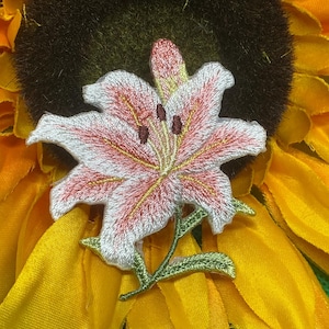 May include: An embroidered patch featuring a lily flower with white and pink petals, yellow stamen details, and dark brown anthers. The flower has a green stem with two leaves extending from the base, showcasing detailed thread work throughout.