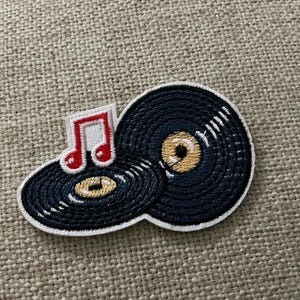 May include: An embroidered patch featuring two black vinyl records with gold centers and a single red musical note symbol. The patch has a white border and is stitched with a textured thread pattern to represent the grooves of the records.