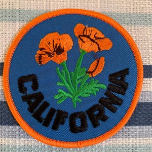 May include: A round patch featuring a blue background with the word "CALIFORNIA" in black lettering. The patch has an orange border and depicts orange California poppies with green stems.