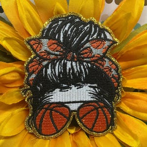 May include: An embroidered patch featuring a black messy bun hairstyle adorned with an orange and black basketball-patterned headband, paired with orange and black basketball-shaped glasses, all outlined with a shimmering gold glitter border.