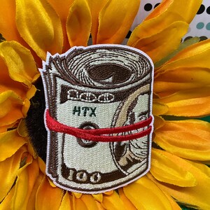 May include: An embroidered patch featuring a rolled-up one hundred dollar bill secured with a red rubber band. The bill design includes the text HTX and the number 100 in brown and cream thread, with a white border surrounding the entire patch shape.