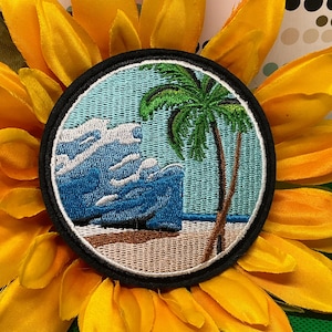 May include: A circular embroidered patch featuring a tropical beach scene with a tall green palm tree, a sandy shore, and a large crashing blue ocean wave. The design is enclosed within a thick black stitched border on a light blue background.
