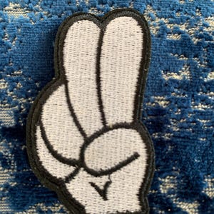 May include: Embroidered patch featuring a white hand in the peace sign gesture, outlined in black. The patch is on a blue textured surface. This patch is suitable for jackets, bags, or other fabric items.