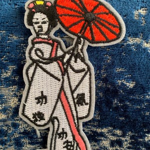 May include: Embroidered iron-on patch featuring a woman in a white kimono holding a red and black umbrella. The kimono has black kanji characters on it.