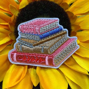 May include: Embroidered patch featuring a stack of books in shades of pink, brown, and blue. The books have detailed stitching and are set against a yellow sunflower background. The patch has a white border.