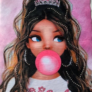 May include: A pink and white graphic tee with a cartoon image of a young woman with brown hair wearing a crown and blowing a pink bubble. The text "Queen" is written in rhinestones on the front of the shirt.