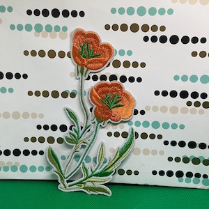 May include: An embroidered patch featuring two vibrant orange poppy flowers with detailed green stems and leaves. The design showcases intricate stitching with a light blue border, highlighting the botanical structure of the flowers and foliage.