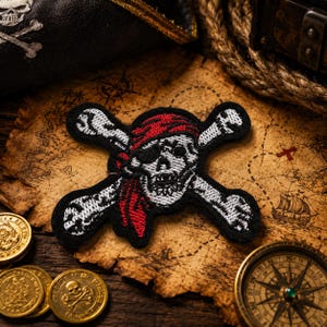 May include: An embroidered patch featuring a white skull wearing a red bandana and an eye patch, positioned over two crossed white bones. The design is outlined in black thread, creating a high-contrast pirate-themed emblem with detailed stitching.