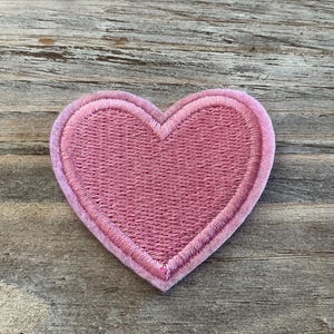 May include: A heart-shaped embroidered patch in a dusty rose pink color. The patch features a dense, textured thread pattern throughout the center and a smooth, raised stitched border that follows the heart shape along the entire outer edge.