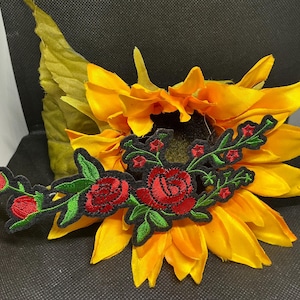 May include: A vibrant yellow sunflower with a black center, adorned with a floral embroidered patch. The patch features red roses, green leaves, and a black border. The sunflower petals are a bright yellow.
