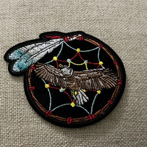 May include: A circular embroidered patch featuring a brown eagle with spread wings centered within a dreamcatcher design. The dreamcatcher has a brown frame, a web with yellow and blue accents, and two white and blue feathers attached to the top.