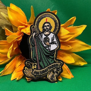 May include: Embroidered patch featuring a religious figure with a halo, holding a staff. The figure is dressed in white and green robes. The patch has the text "SAN JUDAS TADEO" on a banner. The patch is set against a sunflower and green background.