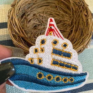 May include: Embroidered patch of a blue and white cruise ship with yellow and red accents. The ship has multiple windows and a detailed design. The patch is on a striped fabric background, with a woven nest in the background.