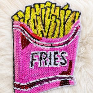 May include: A sequined patch shaped like a container of french fries. The container is bright pink with the word FRIES embroidered in white and black letters. The fries are yellow sequins. The entire patch is outlined in white and black thread.