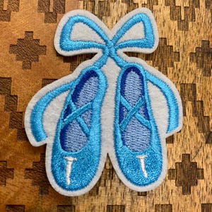 May include: A light blue embroidered patch featuring a pair of ballet slippers with a bow. The patch has a white felt backing and blue embroidered details. The ballet slippers are a vibrant blue color.