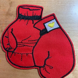 May include: A red embroidered patch featuring two boxing gloves. The gloves are red with black outlines and details. One glove has a white and yellow accent. The patch is designed for application on clothing or accessories.