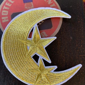 May include: Embroidered patch featuring a gold crescent moon and two stars, outlined in white. The patch has a detailed, textured appearance. The background includes a vintage Hotel Continent label.
