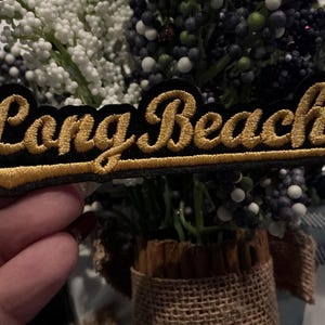 May include: A black embroidered patch with the words "Long Beach" in gold script. The patch is rectangular and has a gold border. The patch is held in front of a floral arrangement.