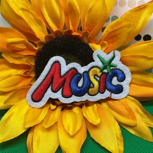 May include: An embroidered patch featuring the word Music in a stylized font with red, blue, and yellow letters on a white background, accented by a small green star above the letter i, resting on a large yellow sunflower with a dark brown center.