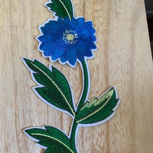 May include: Embroidered floral patch featuring a vibrant blue flower with a yellow center and green leaves. The patch has a white border and is designed to be sewn or ironed onto fabric. Ideal for adding a decorative touch to clothing or accessories.