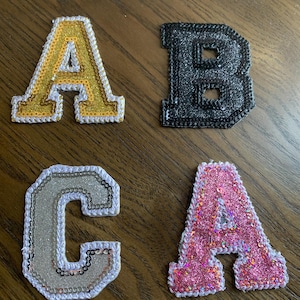 May include: Four embroidered fabric letters: gold A, black B, silver C, and pink A. Each letter is outlined with sequins. The letters are likely for personalization on clothing or accessories.