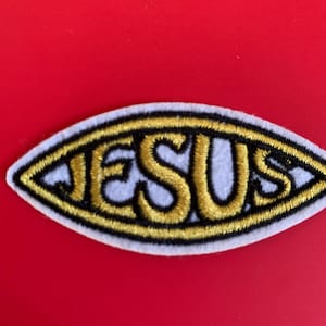 May include: Embroidered patch in an oval shape with the word "JESUS" in gold thread. The patch has a white background with a black and gold border. The background is a solid red color.