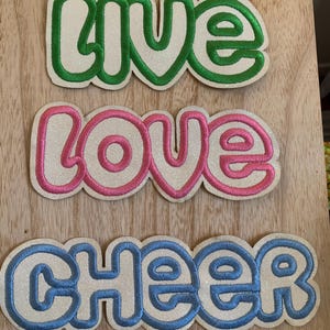 May include: Three embroidered patches with the words "LIVE", "LOVE", and "CHEER". The "LIVE" patch is green, the "LOVE" patch is pink, and the "CHEER" patch is blue. Each patch has a glittery, off-white background.