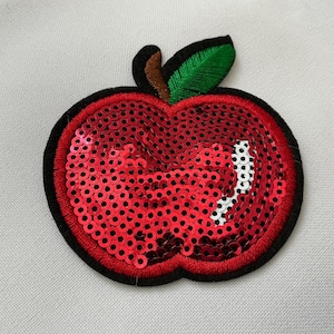 May include: A red sequined apple patch with a black border, a brown stem, and a green leaf. The apple's surface is covered in small, shiny red sequins. This decorative patch can be ironed on to clothing or accessories.