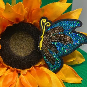May include: A close-up of a bright yellow sunflower with a dark brown center. A sequined butterfly with blue and brown wings is attached to the flower. The butterfly has a black outline and yellow antennae.