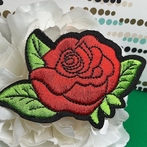 May include: An embroidered patch featuring a vibrant red rose with detailed black stitching for the petals and bright green leaves. The entire design is outlined with a thick black border, creating a bold contrast against the floral embroidery.