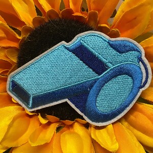 May include: An embroidered patch shaped like a whistle, featuring shades of light blue, medium blue, and dark blue thread with a white border. The design depicts a classic whistle structure with a mouthpiece and a circular ring attachment point.