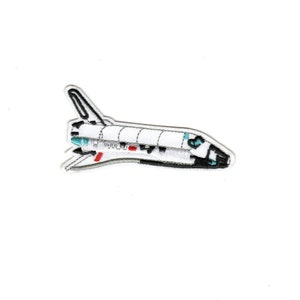May include: White and black embroidered patch of a space shuttle in flight. The shuttle has a red stripe and a blue and white design on the nose.