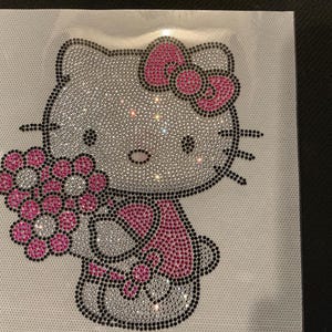 May include: A rhinestone-studded Hello Kitty design, holding a bouquet of flowers. The design features a pink bow, dress, and flowers, with black outlines and accents. The rhinestones are silver.