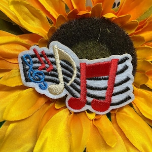 May include: An embroidered patch featuring a white background with black horizontal musical staff lines. The design includes a blue treble clef, a red eighth note, a gold-colored musical note, and a large red double eighth note symbol.