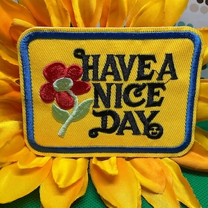 May include: A rectangular embroidered patch with a yellow background and a blue border. The text "HAVE A NICE DAY" is stitched in black, with a red flower featuring a light green stem and leaves positioned to the left of the lettering.