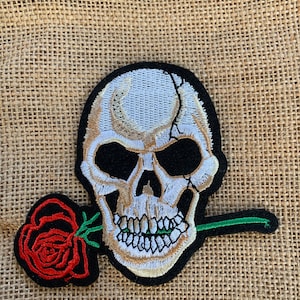 May include: Embroidered iron-on patch featuring a white skull with black eyes and a red rose in its mouth. The skull is holding a green stem in its teeth.