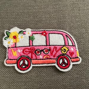May include: Embroidered patch featuring a pink van with the word "GROOVY" in red. The van has white windows, red wheels, and is decorated with flowers. The patch is outlined in white and is likely for clothing or accessories.