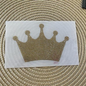 May include: A gold crown-shaped iron-on patch with rhinestones. The patch is on a white fabric background.